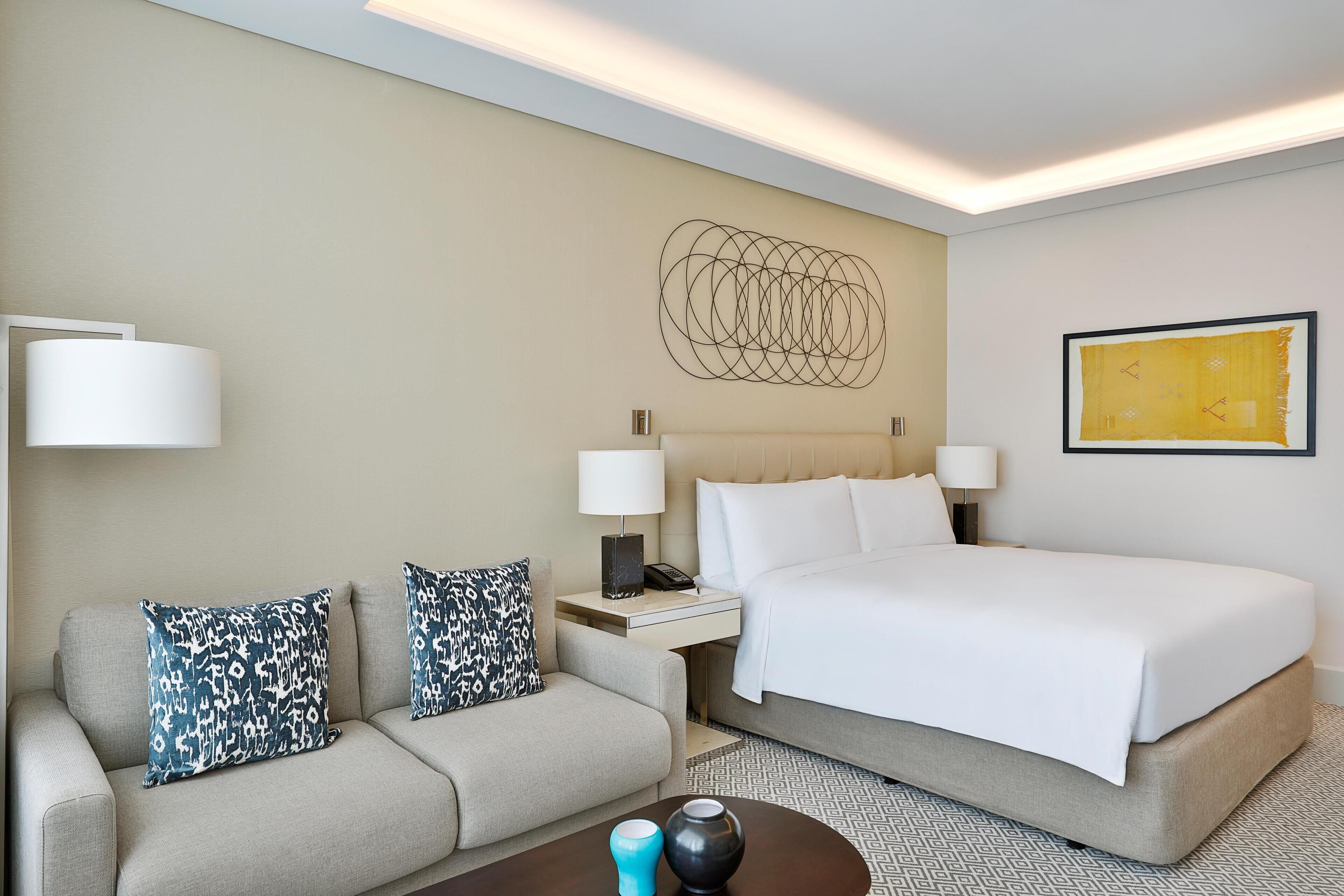 A hotel room with a bed, a sofa, two lamps, a nightstand, and a wall art piece.