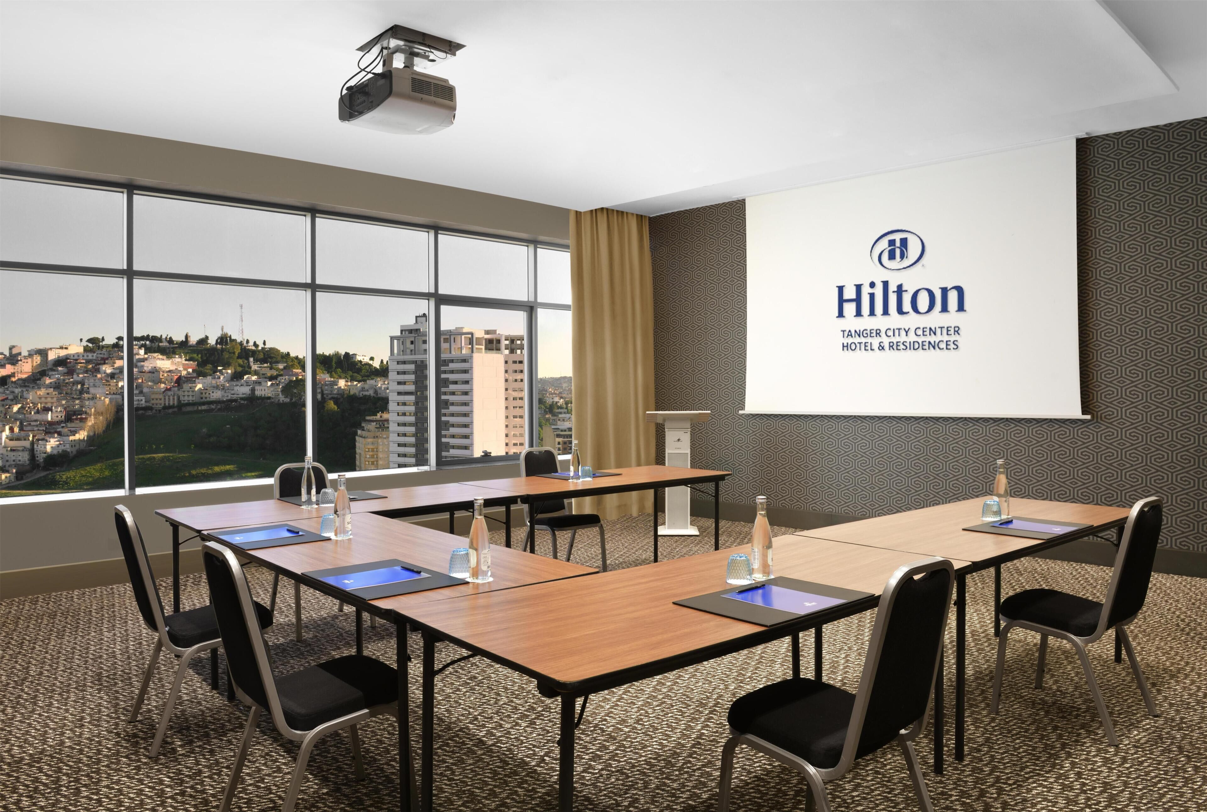 A conference room with a large screen displaying the Hilton logo, a city view, and a carpeted floor.