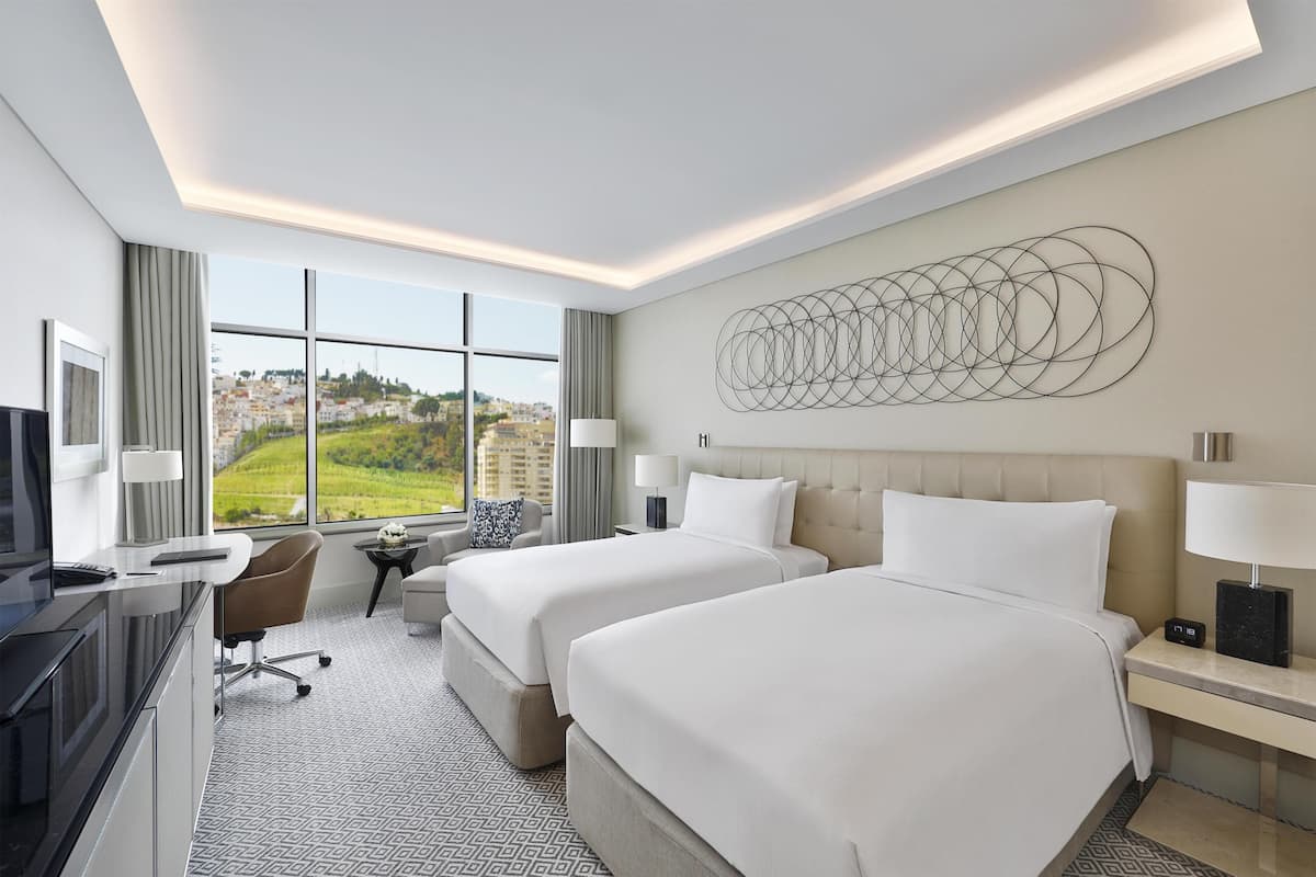 A hotel room with two beds, a desk, a TV, and a large window with a city view.