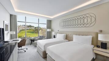 Habitación de hotel con dos camas, un escritorio, televisor y un amplio ventanal con vistas a la ciudad.