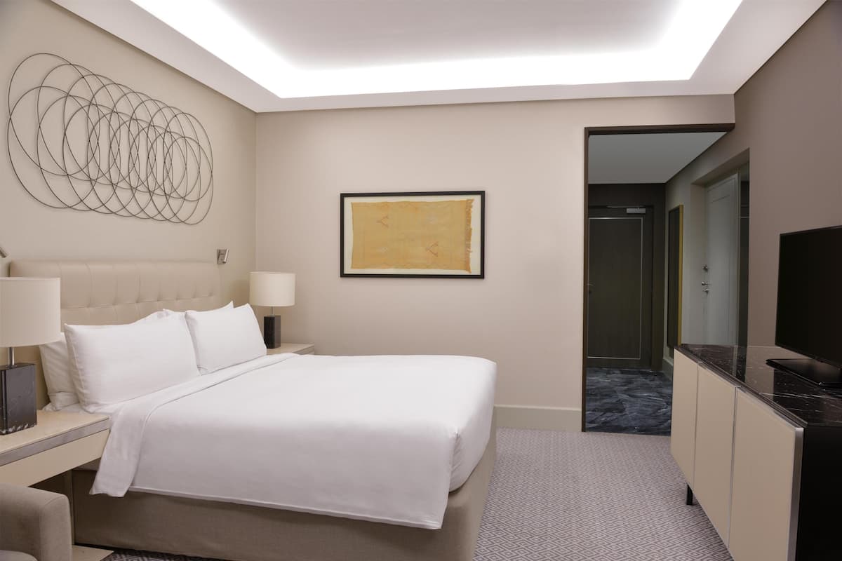 A hotel room with a bed, bedside tables, a TV, a framed artwork, and a door leading to a bathroom.