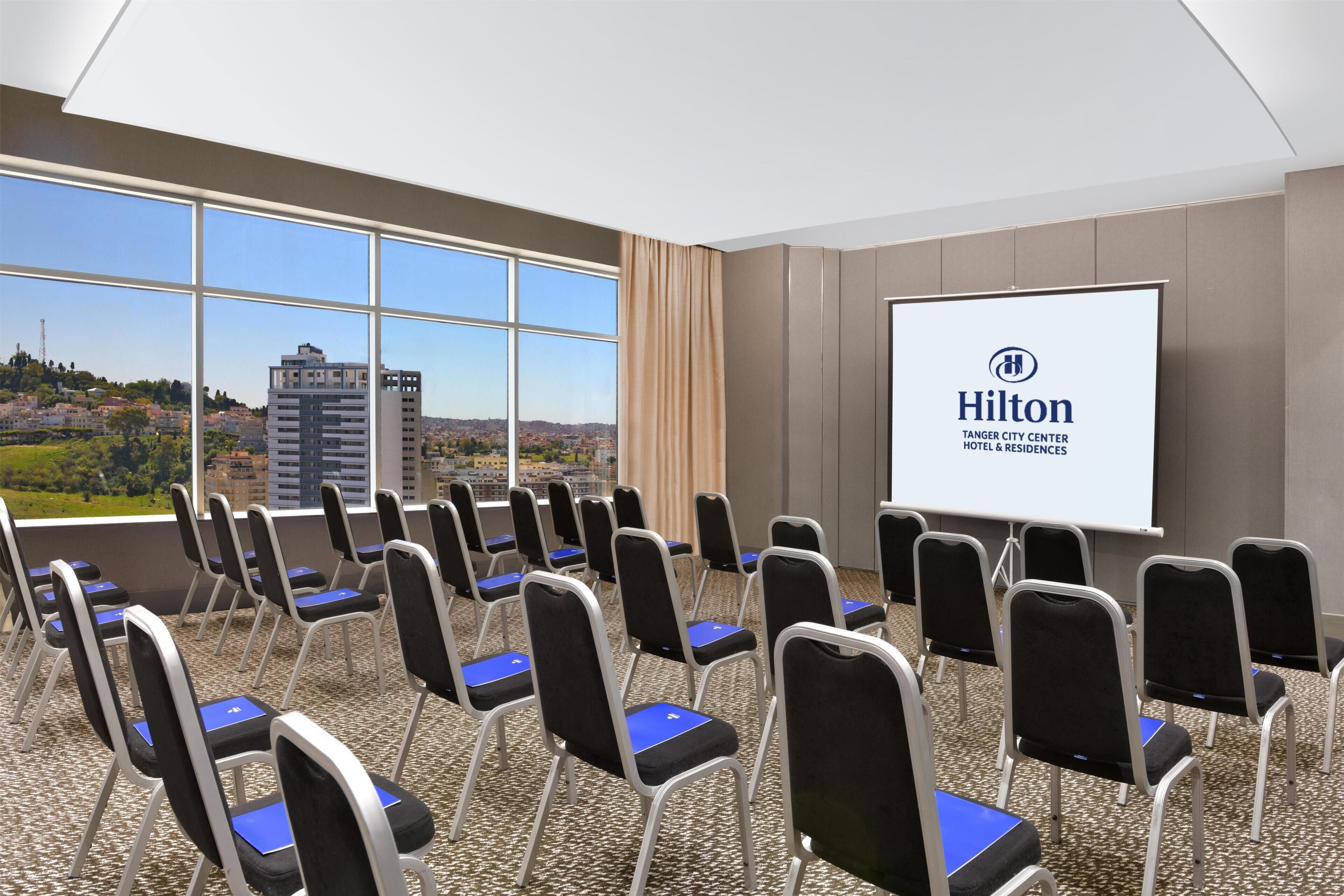 Une salle de conférence avec des rangées de chaises, un écran de projection affichant le logo de Hilton et de grandes fenêtres donnant sur le paysage urbain.