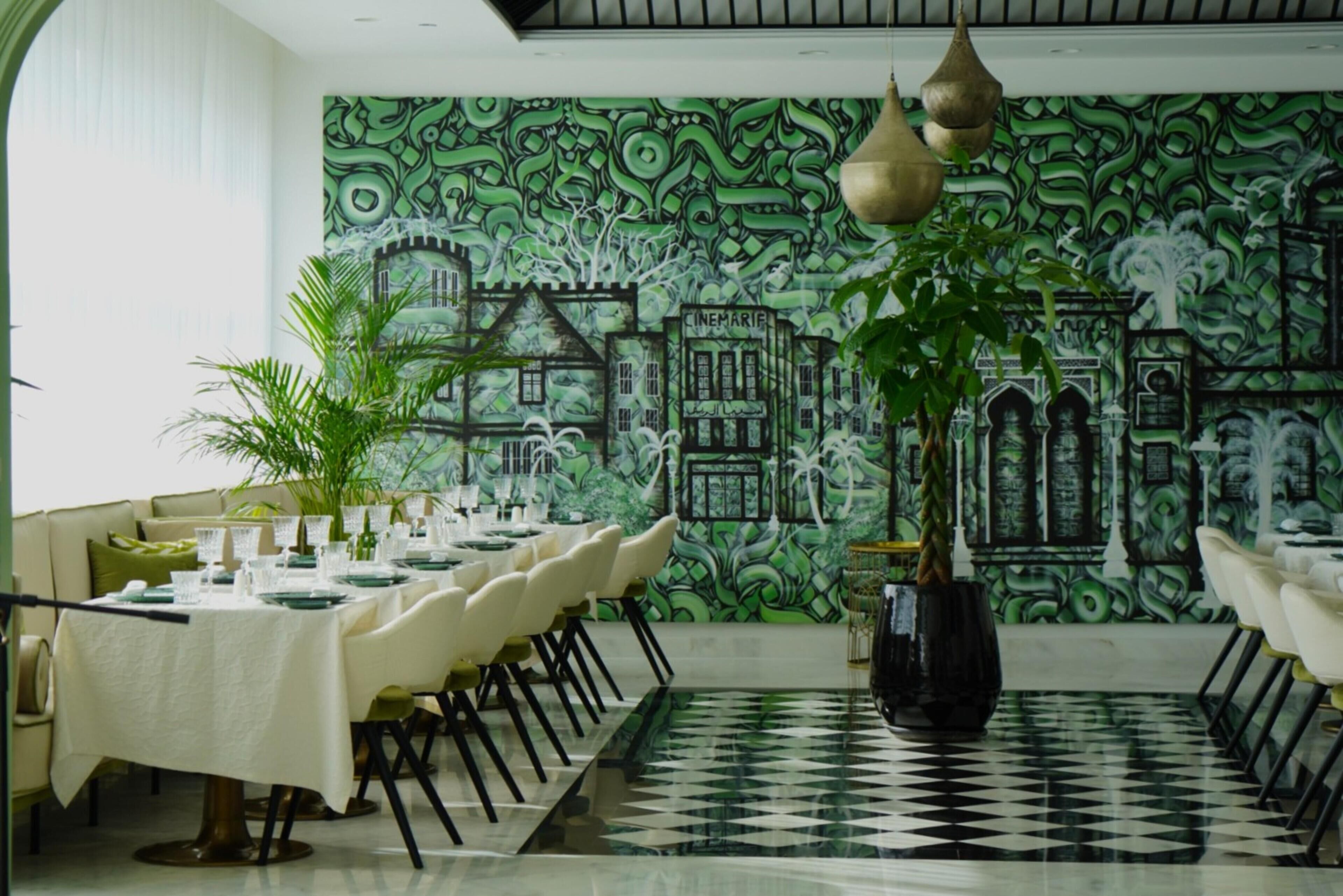 A dining area with a long table set for a meal, a patterned wall mural, and a potted plant.