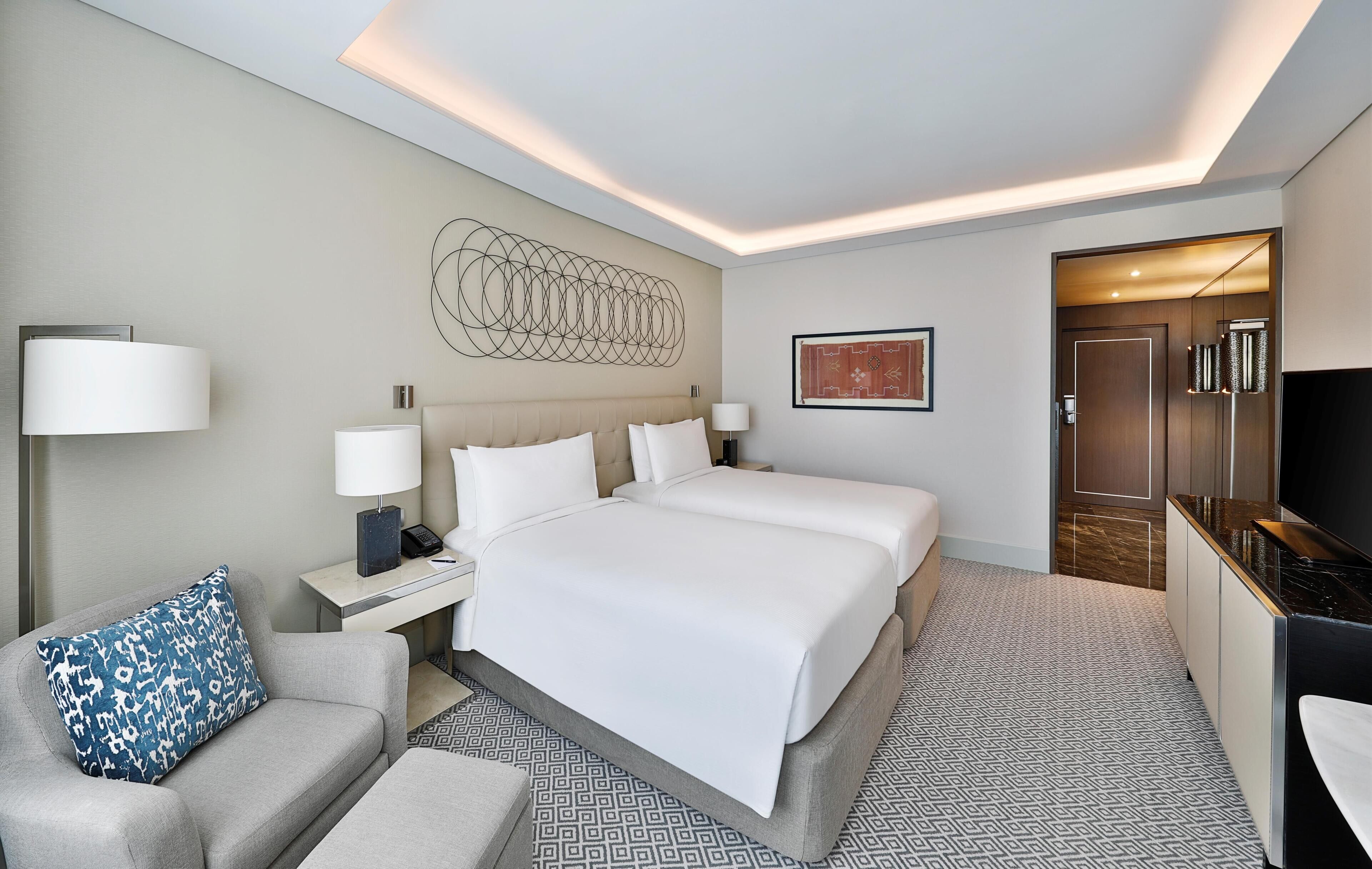 A hotel room with a large bed, bedside tables, a desk, a chair, a TV, and a door leading to another room.