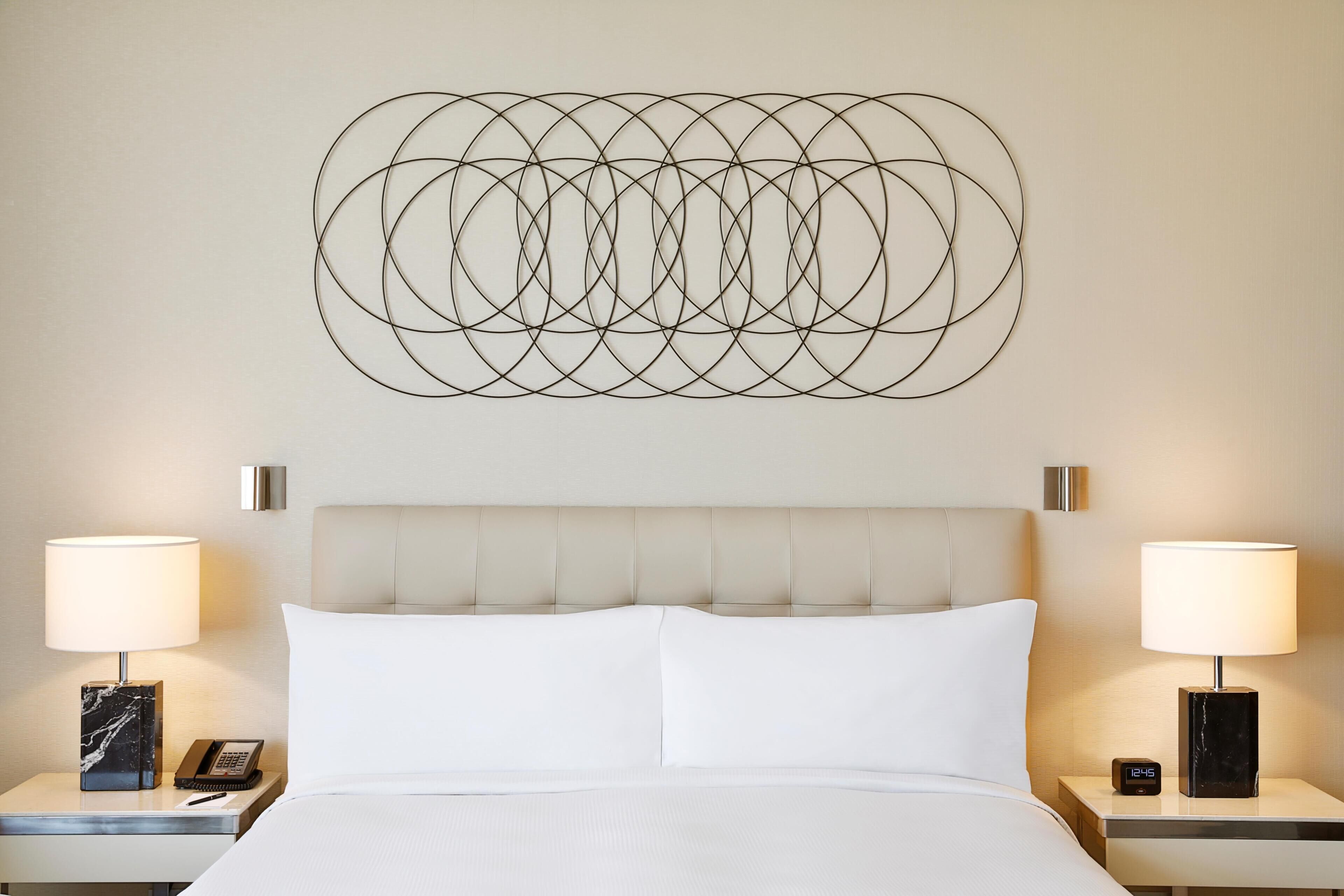 Une chambre d’hôtel comprenant un lit, deux tables de chevet avec des lampes, une horloge murale et une œuvre d’art géométrique accrochée au mur.