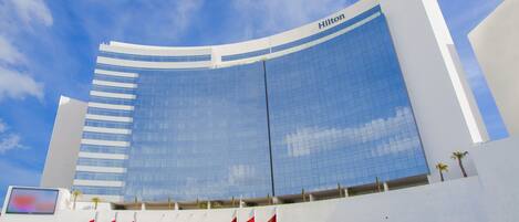 Un hotel Hilton moderno con fachada de vidrio curva, múltiples mástiles de banderas y cielo despejado.
