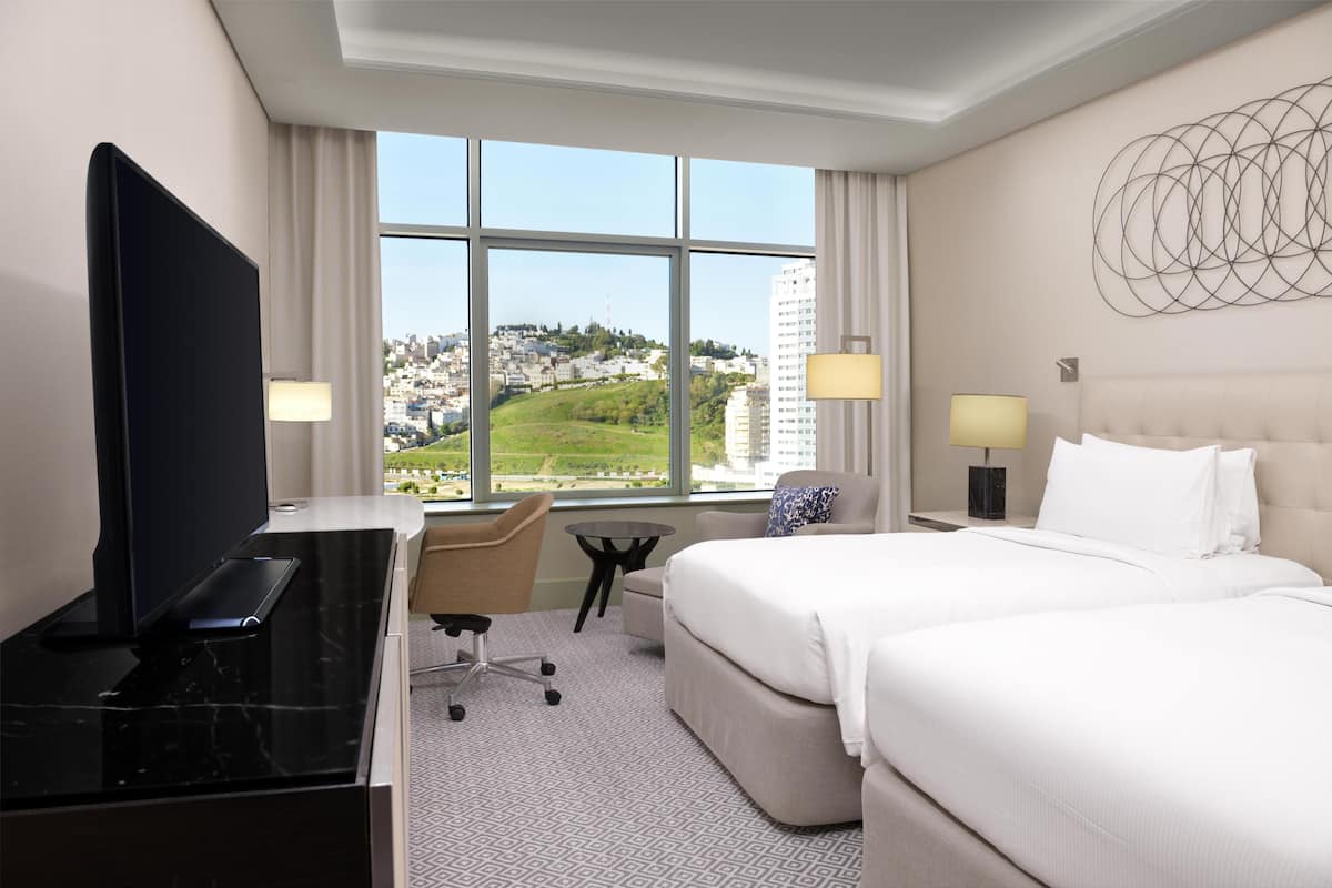 A hotel room with a bed, a desk with a TV, a chair, a small table, and a view of a cityscape.