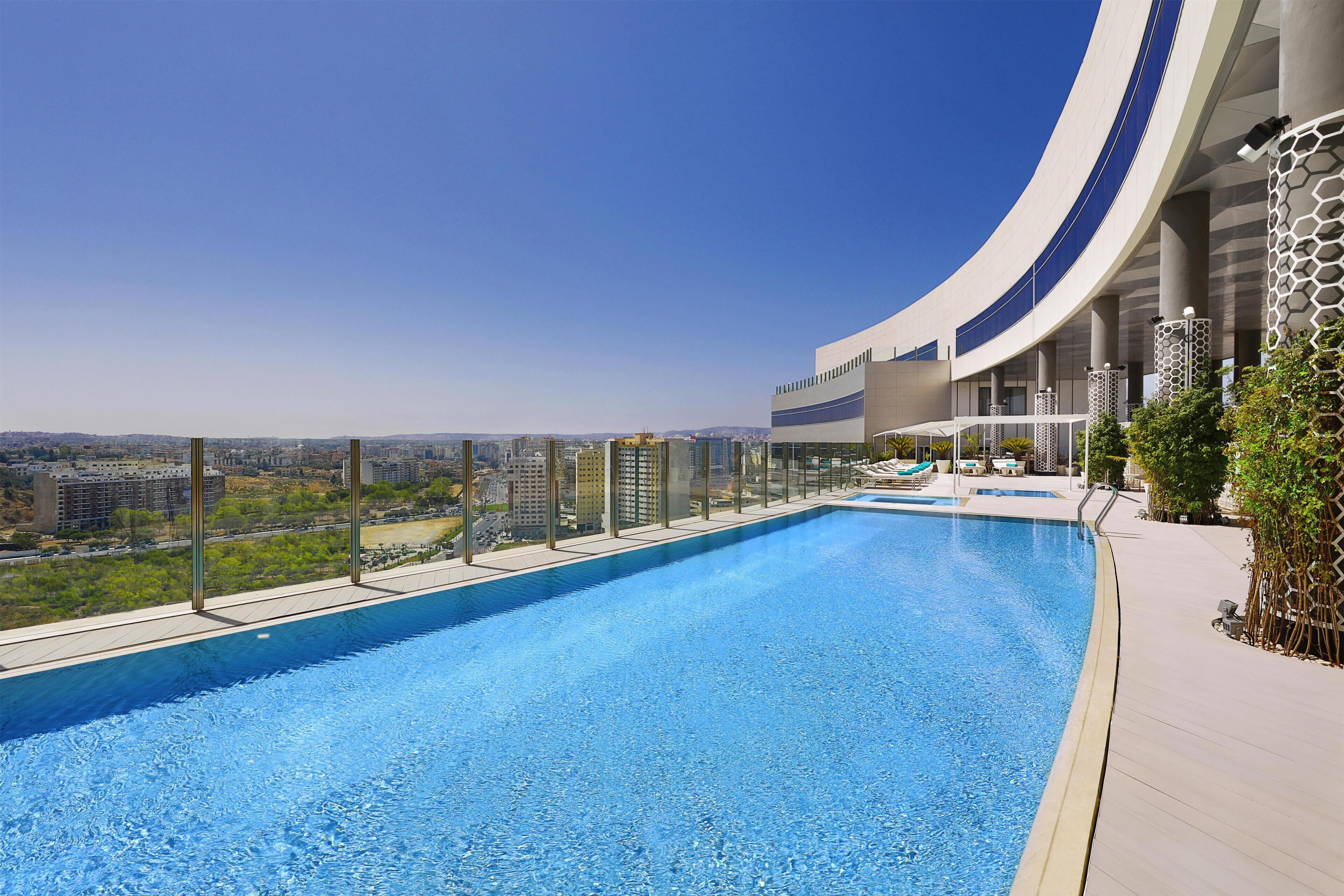 A rooftop infinity pool with a city view, a curved building structure, and a clear sky.
