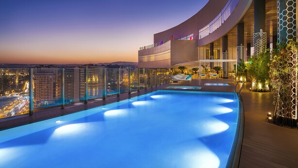 A rooftop infinity pool with a city view, a curved building with large windows, and a wooden deck with lounge chairs.