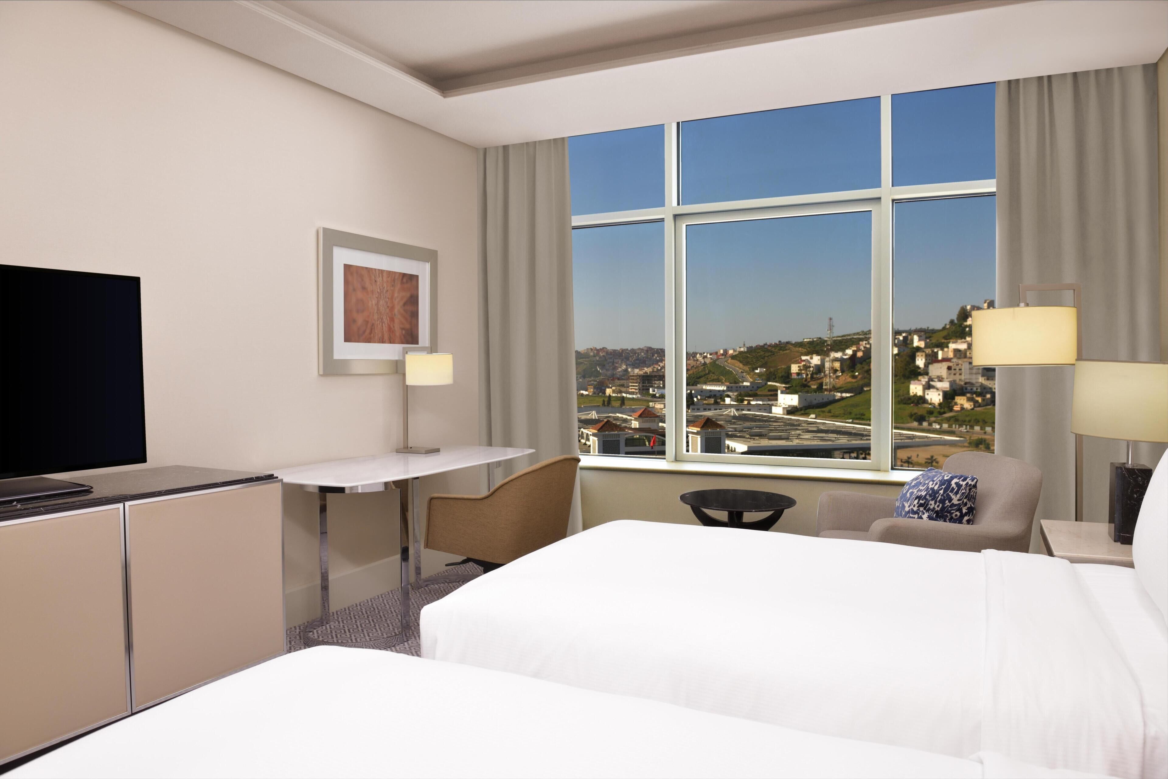 Ein Hotelzimmer mit Bett, Schreibtisch, Fernseher und einem Fenster mit Blick auf die Stadt.