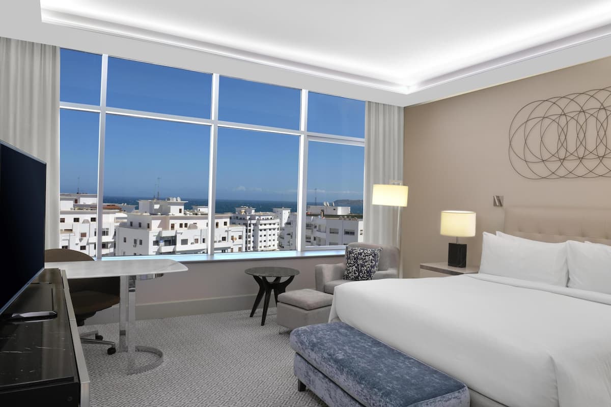 A hotel room with a large window, a desk, a chair, a bed, and a nightstand with a lamp.