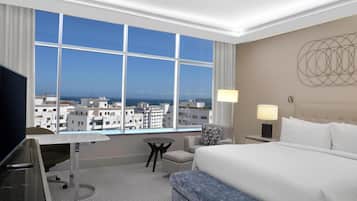 Une chambre d’hôtel avec une grande fenêtre, un bureau, une chaise, un lit et une table de chevet avec une lampe.
