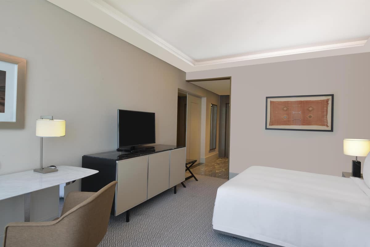 A hotel room with a bed, desk, chair, television, and artwork on the wall.