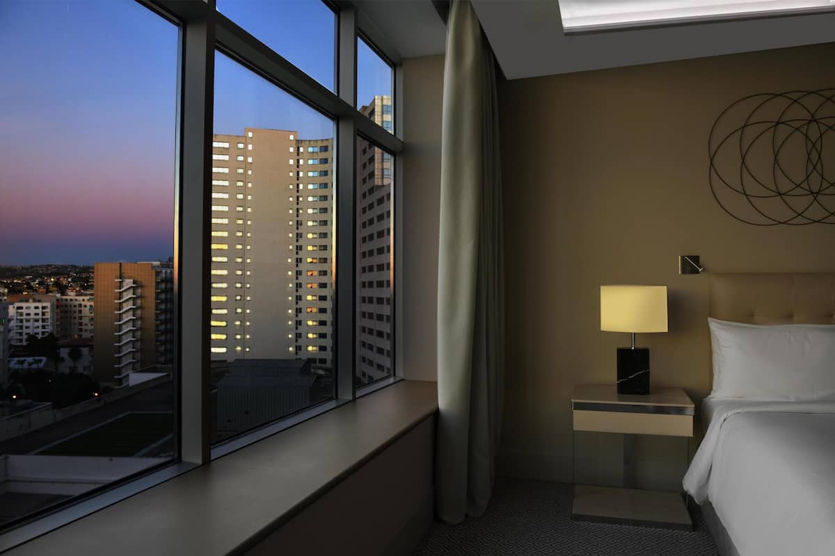 A hotel room with a large window, a bedside table with a lamp, a bed, and a wall art piece.