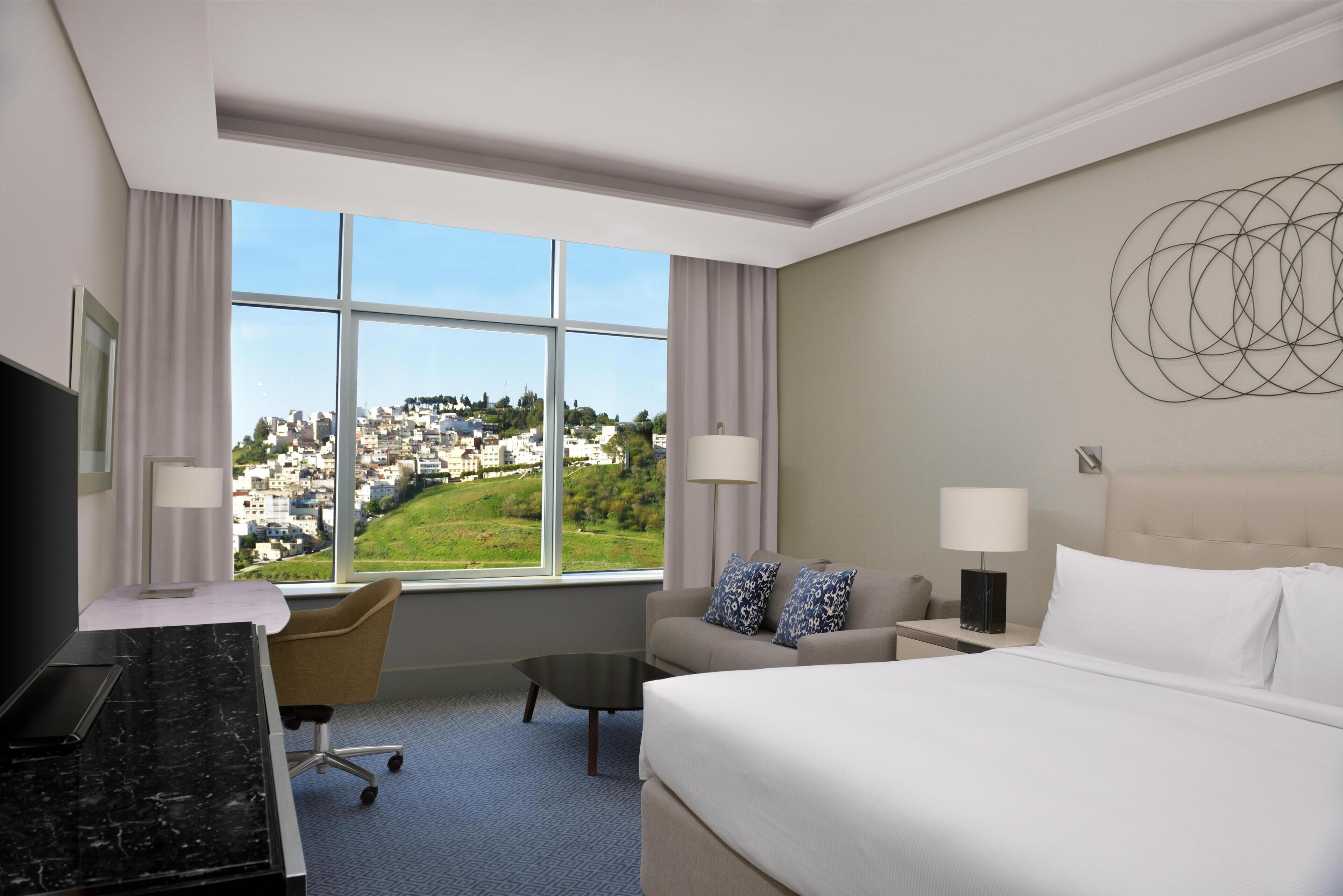 A hotel room with a bed, a desk, a chair, a nightstand, and a view of a cityscape through large windows.