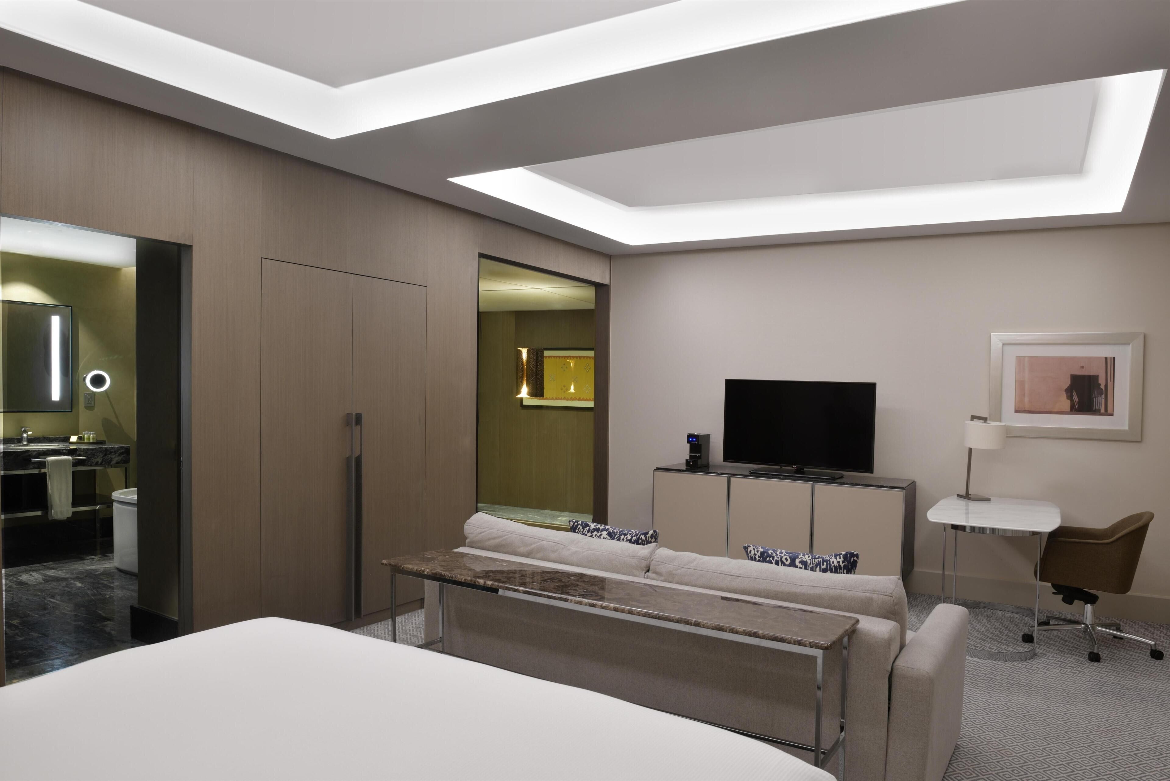 A modern hotel room with a bed, a TV, a desk, and a bathroom visible through an open door.