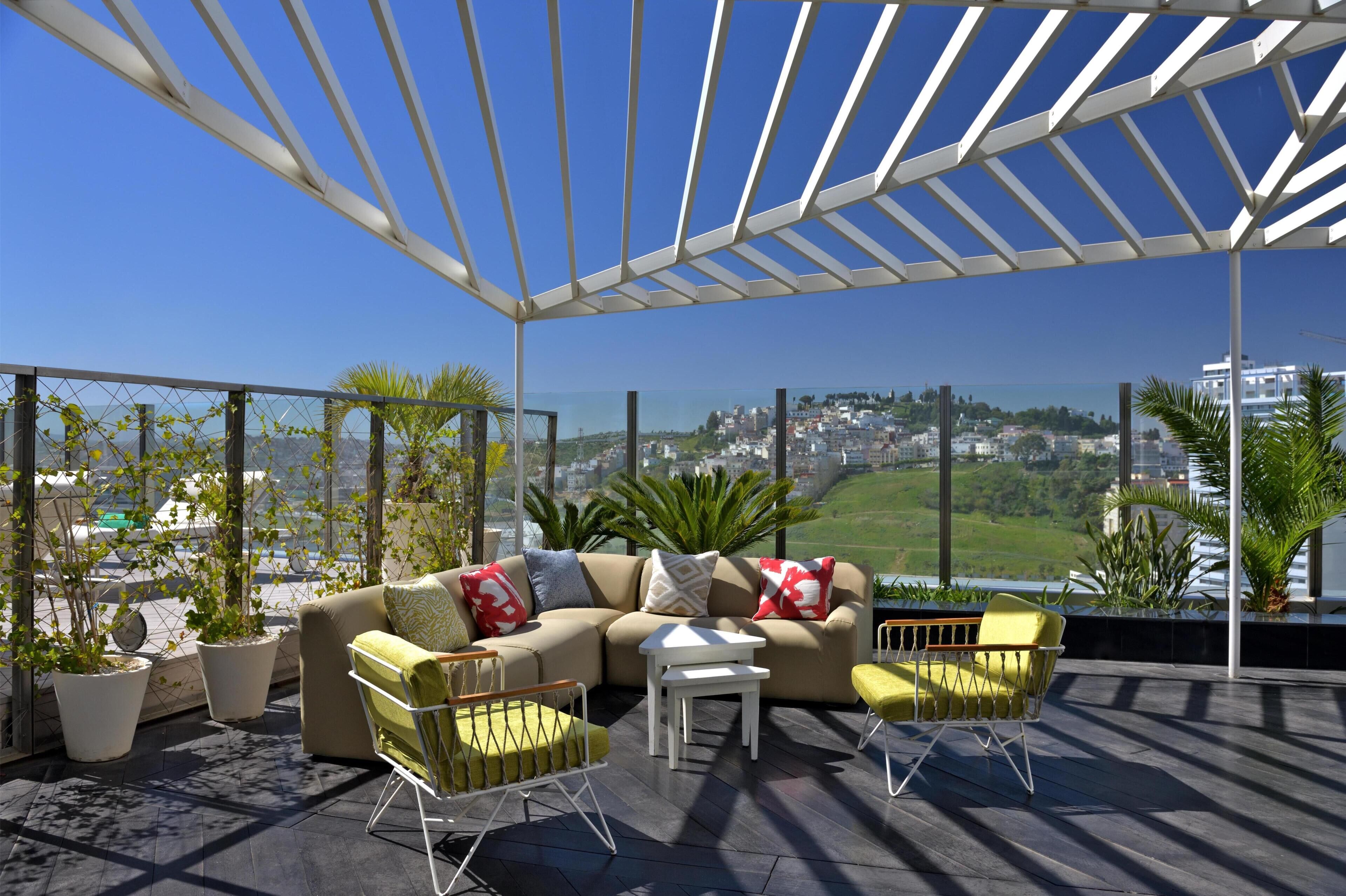 Una terraza en la azotea con una pérgola blanca, un sofá color crema con cojines, dos sillas amarillas y plantas en macetas.