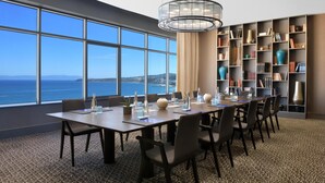 A conference room with a large table, chairs, a bookshelf, and a view of the sea.