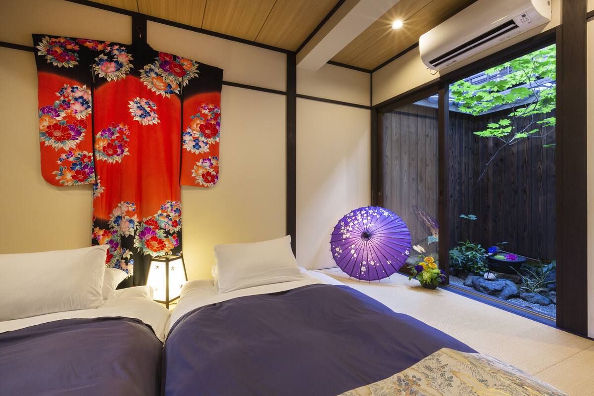 A hotel room with a bed, a traditional Japanese folding screen, a purple umbrella, and a view of a garden through a sliding door.