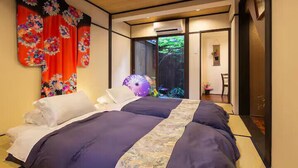 Una camera da letto tradizionale giapponese con tatami, un letto con biancheria scura, uno schermo scorrevole che dà su un giardino e un arazzo floreale dai colori vivaci appeso alla parete.