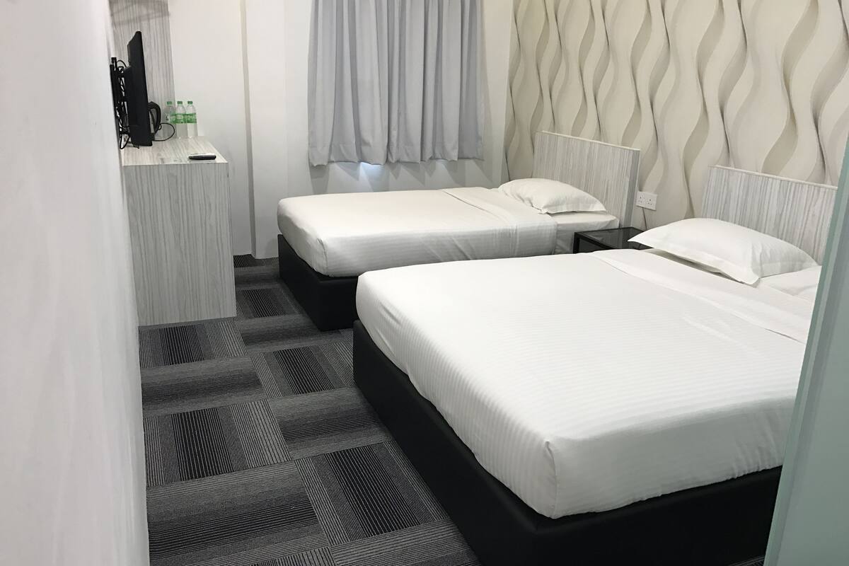 Ett hotellrum med två sängar, ett litet skrivbord med en tv och några flaskor, grå gardiner och en vägg med ett mönster.