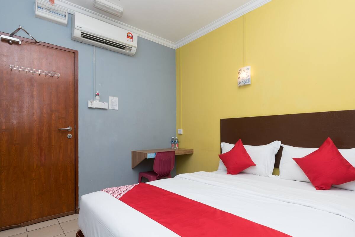 A hotel room with a wooden door, a bed with white and red bedding, a bedside table with a red chair, and an air conditioning unit on the wall.