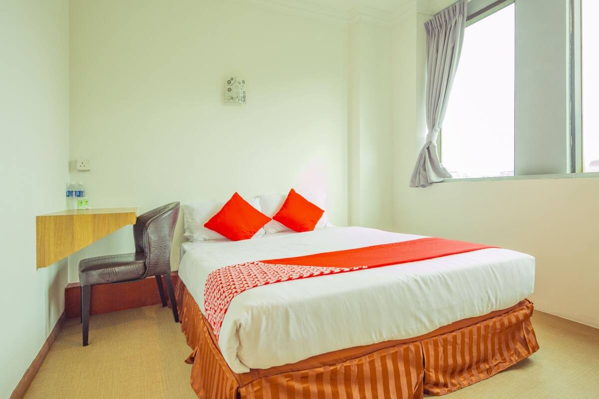 A hotel room with a bed, a desk, a chair, a window with curtains, and a wall socket.