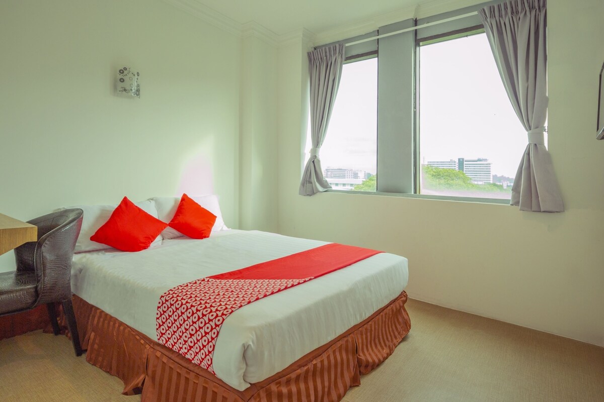 A hotel room with a bed, a chair, a window with curtains, and a view of buildings outside.