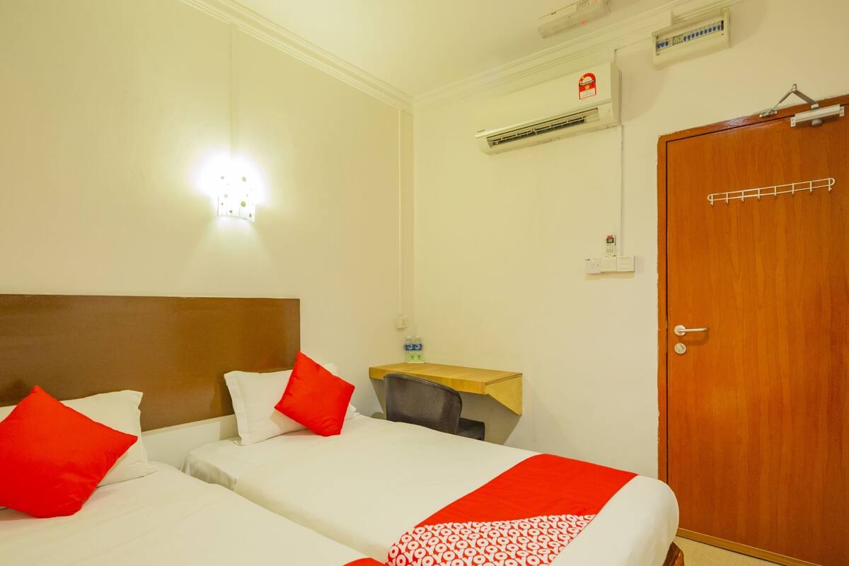 A hotel room with two beds, each with white bedding and red accents, a wooden headboard, a small desk with a chair, a wooden door, and an air conditioning unit on the wall.