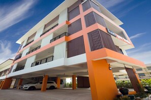 A multi-story hotel with a modern design, featuring balconies on each floor and a parking area with cars.