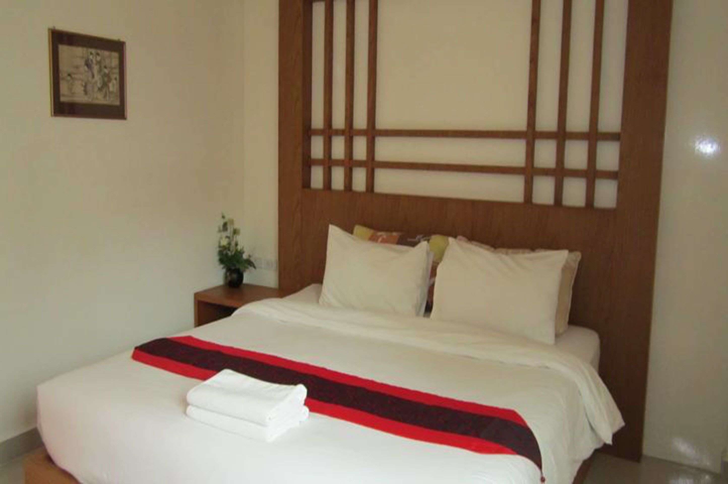 A neatly made bed with white bedding and a red and black striped blanket, a wooden headboard with a grid pattern, and a small bedside table with a plant.