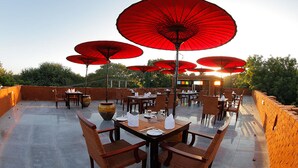 An outdoor dining area with multiple tables and red umbrellas, surrounded by brick walls and greenery.