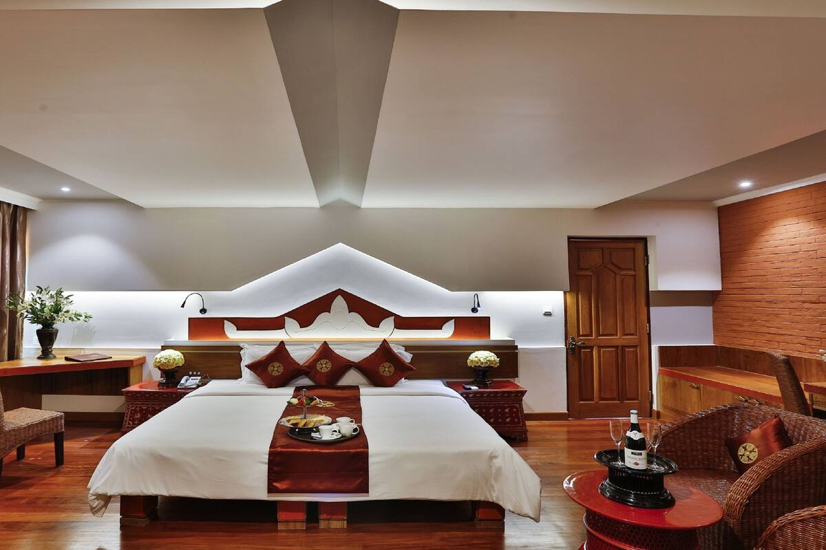 A hotel room with a large bed, a wooden headboard, a dining table with a red table runner, wicker chairs, and a wooden door.