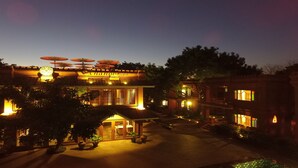 A hotel with a central building featuring a covered outdoor seating area with umbrellas, surrounded by trees and a well-lit courtyard.