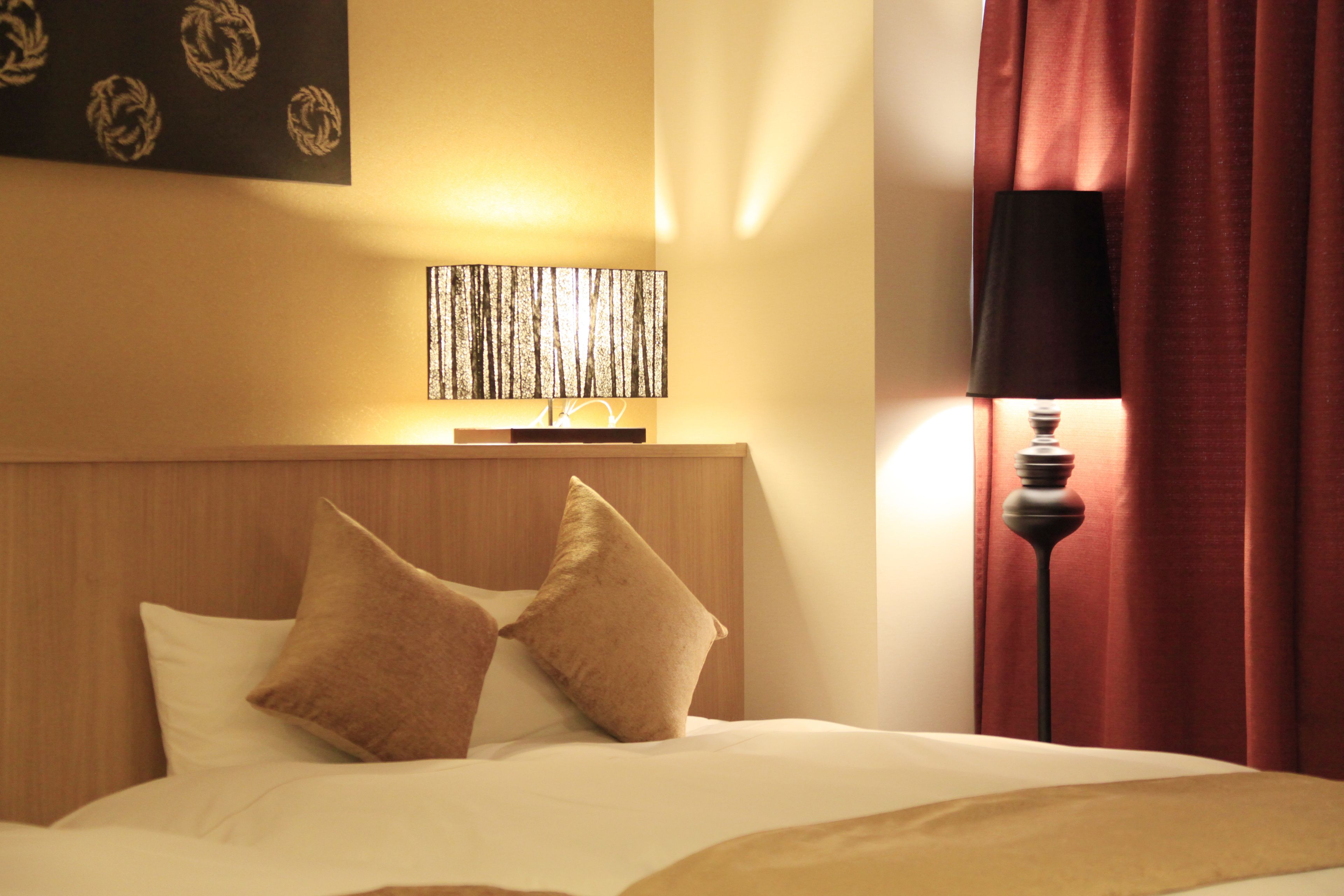 A hotel room with a bed, two pillows, a wall-mounted lamp, and a red curtain.