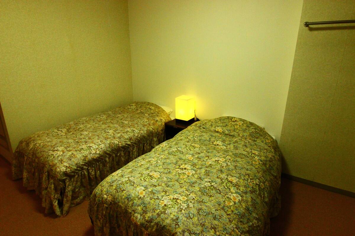 A small room with two single beds, a nightstand with a lamp, and a towel rack on the wall.