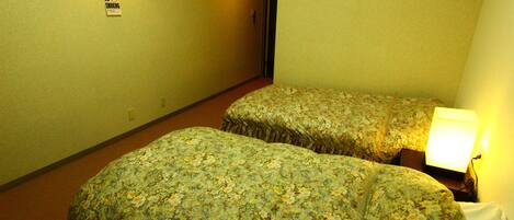 A hotel room with two single beds, a nightstand with a lamp, a wall-mounted light, and a 'No Smoking' sign.