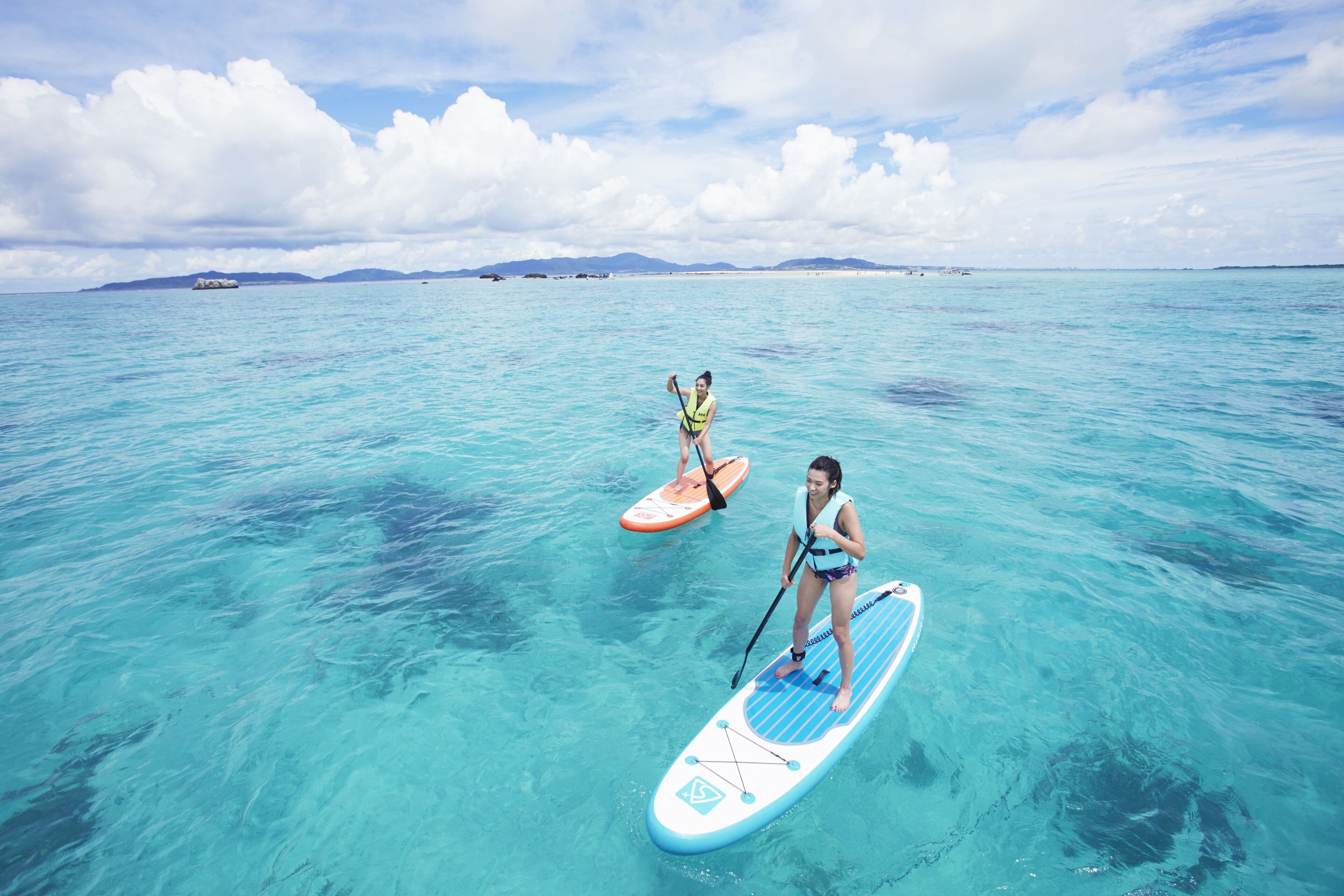 Two people paddleboarding in clear turquoise water with distant land and boats visible.