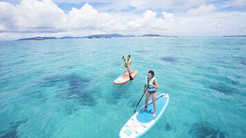 Two people paddleboarding in clear turquoise water with distant land and boats visible.