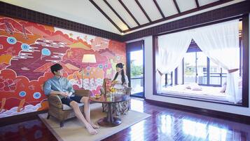 A room with a large window, a colorful mural, wicker furniture, and a small table with a lamp.