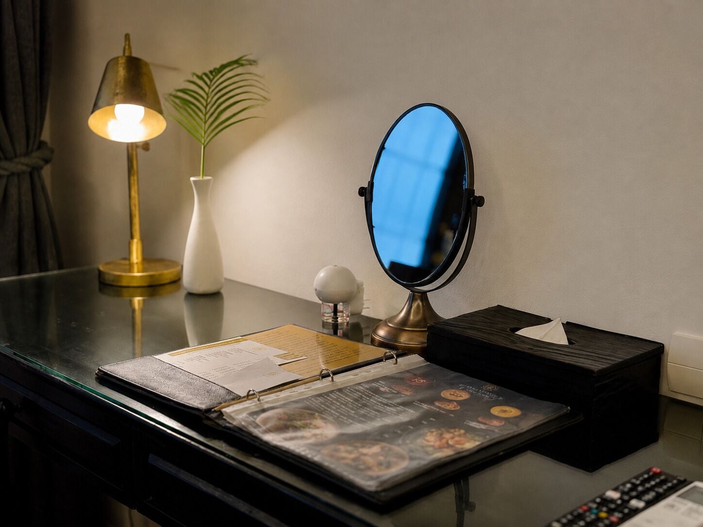 A hotel room with a desk, a lamp, a mirror, a vase with a plant, and a menu.