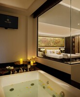 A modern bathroom with a large bathtub, a mirror reflecting a bedroom, and a TV mounted on the wall.
