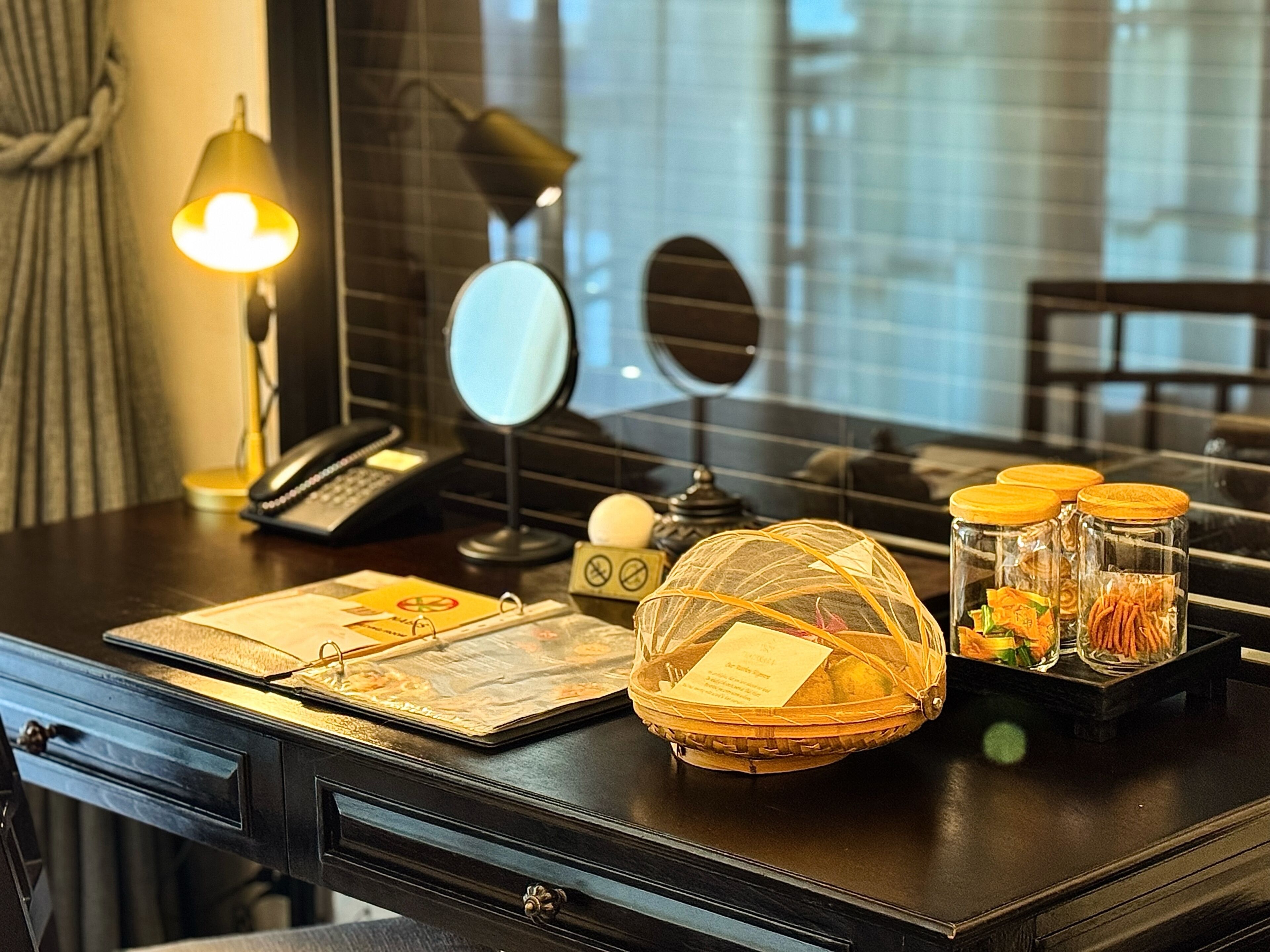 A hotel room desk with a lamp, phone, mirror, and decorative items including a basket and jars.