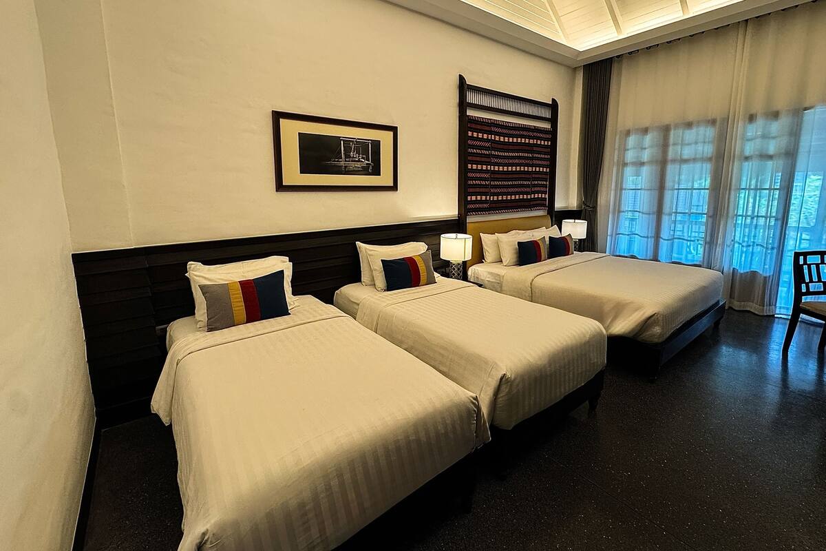 A hotel room with three beds, a large window with curtains, a framed picture on the wall, and a ceiling with a skylight.