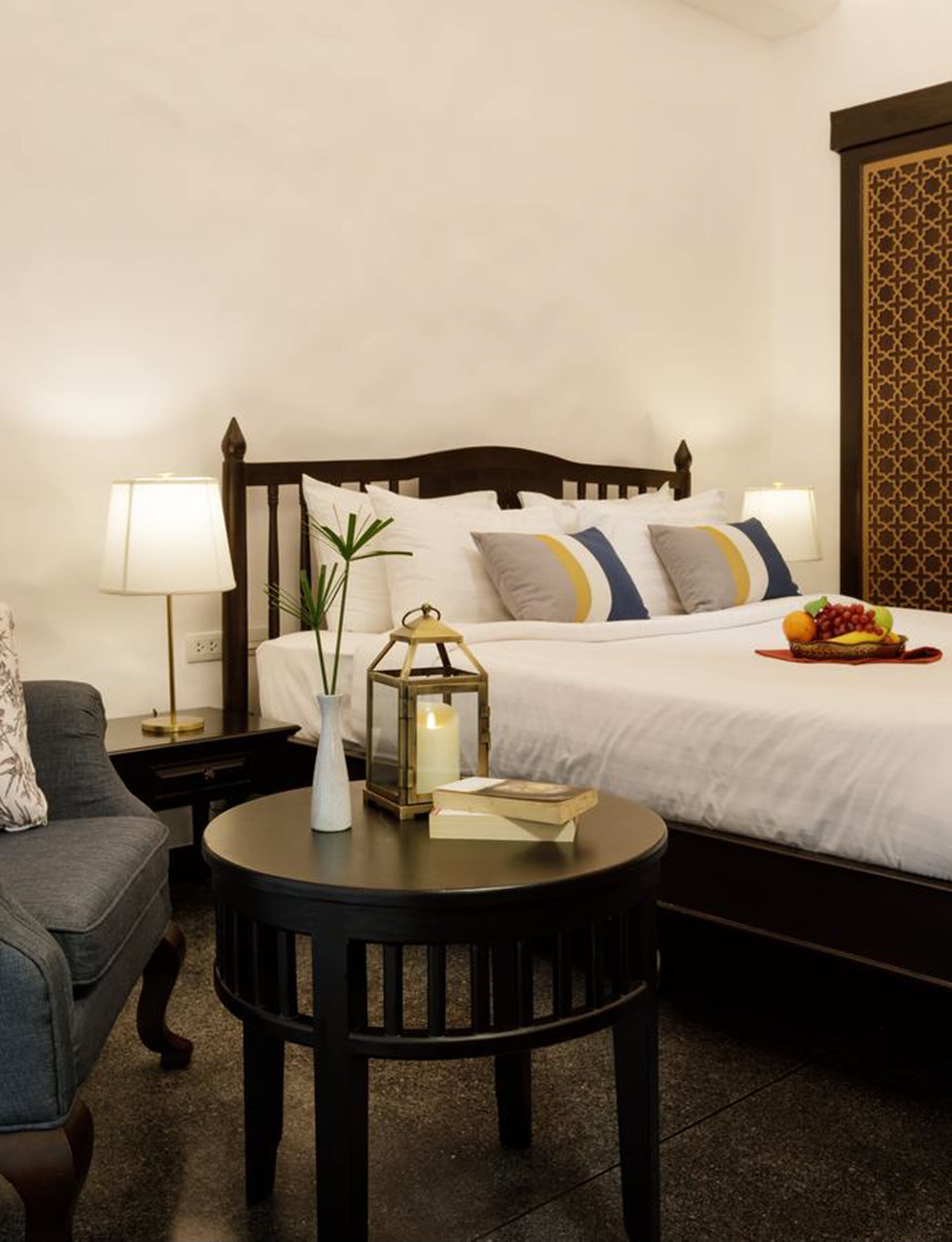 Habitación de hotel ordenada con cama, lámparas de noche, una mesita redonda con un libro y una vela, y una cesta de frutas sobre la cama.