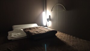 A dimly lit room with a bed, a wall-mounted lamp, and a folded towel on the bed. - Book Tea Bed GINZA - Hostel (Tokyo)
