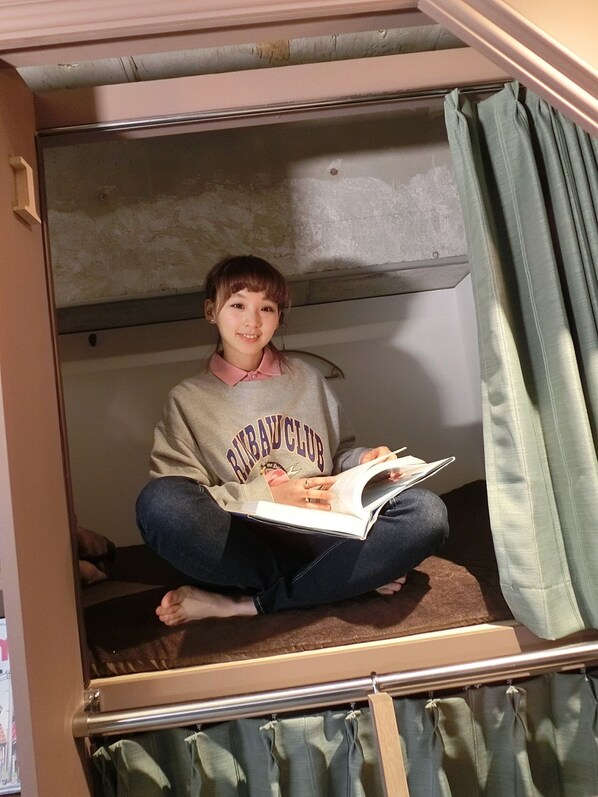 A person sitting on a bunk bed in a room with a bunk bed, a window with curtains, and a visible door. - Book Tea Bed GINZA - Hostel (Tokyo)