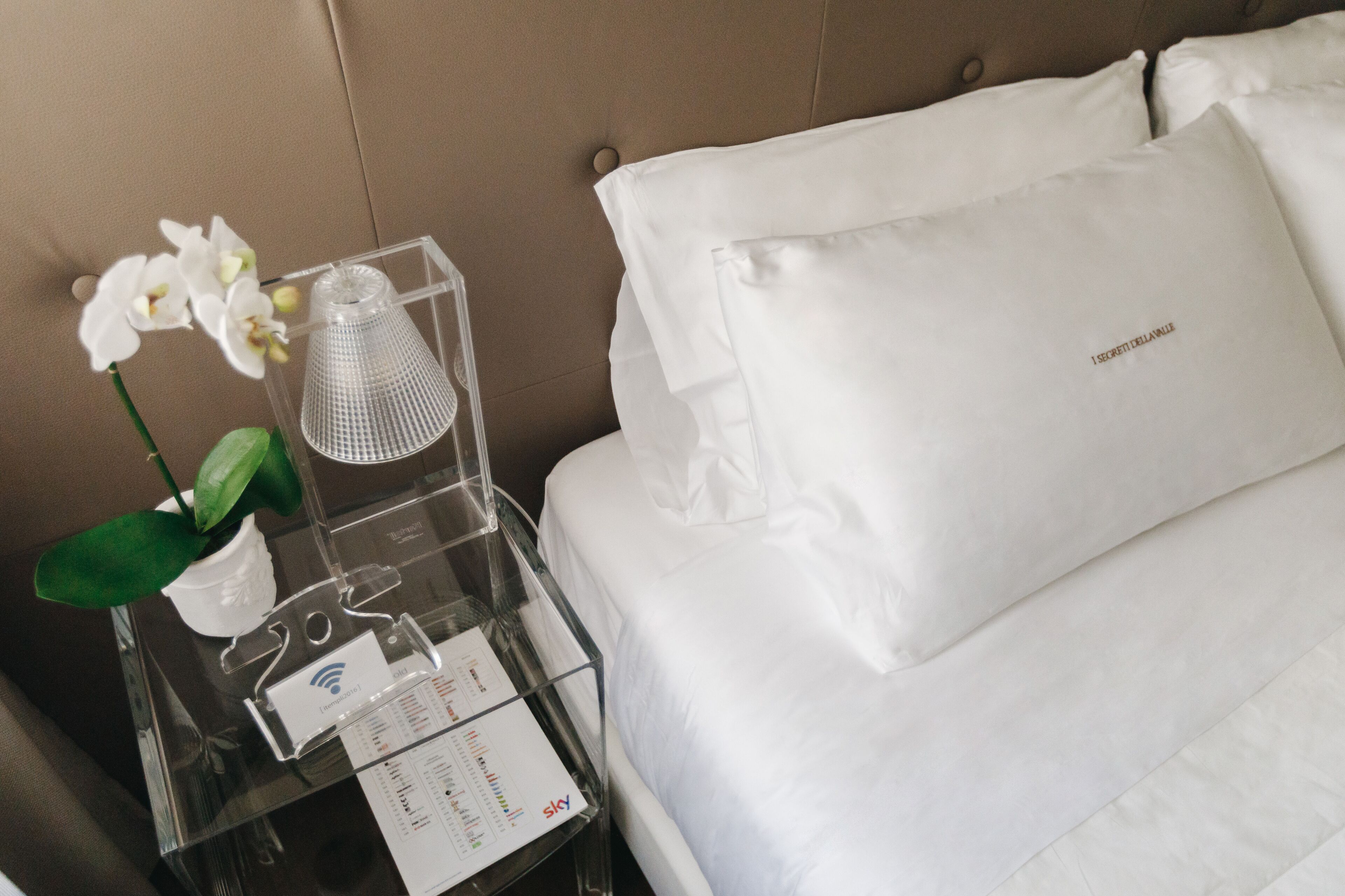A neatly made hotel bed with white linens and a pillow with 'I SECRET DELANNE' embroidered on it.