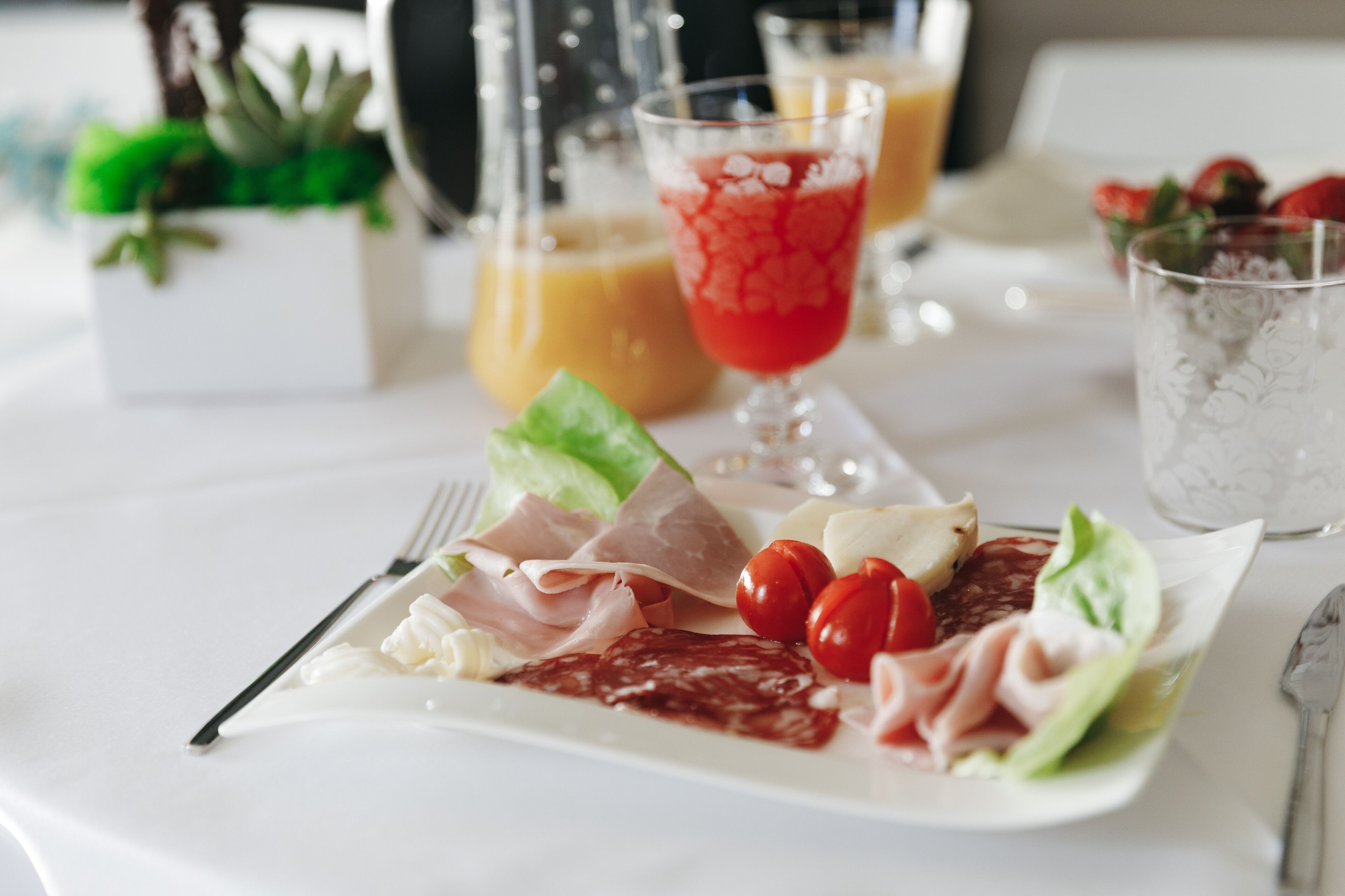 Una mesa blanca con un plato de comida, que incluye carnes, queso y tomates, y dos vasos de jugo.