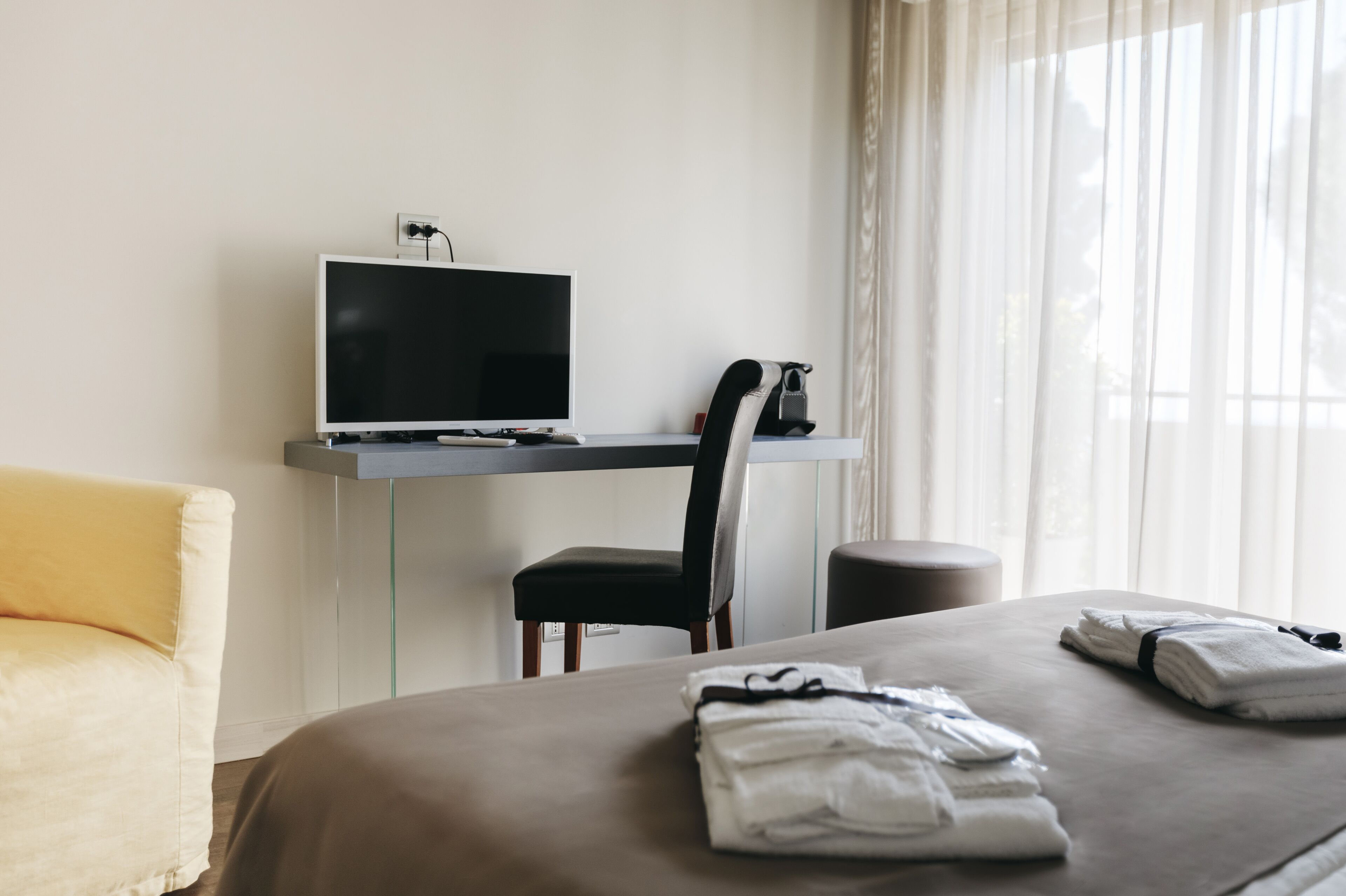 A hotel room with a bed, a desk with a computer, a chair, and a window with curtains.